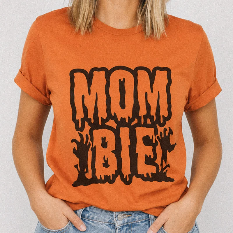 Woman wearing a burnt orange MomBie Funny Zombie Mom Halloween Tee featuring bold dripping text that reads Mombie
