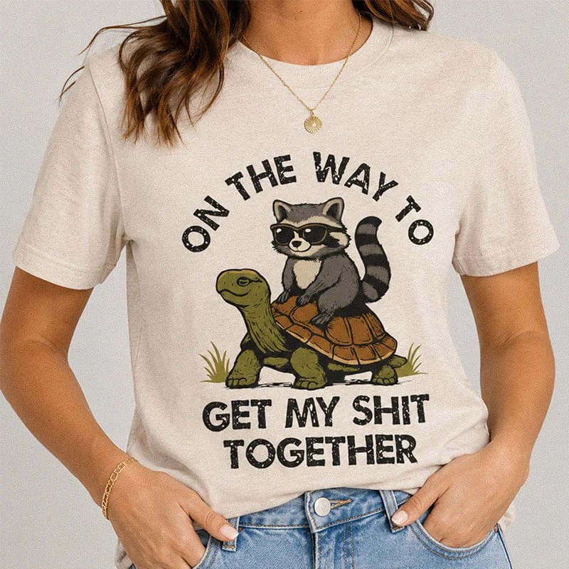 Woman wearing a heather dust On the Way to Get My Shit Together Shirt - Funny Procrastination Shirt featuring a sunglasses-wearing raccoon riding a slow turtle with the text "On The Way To Get My Shit Together"