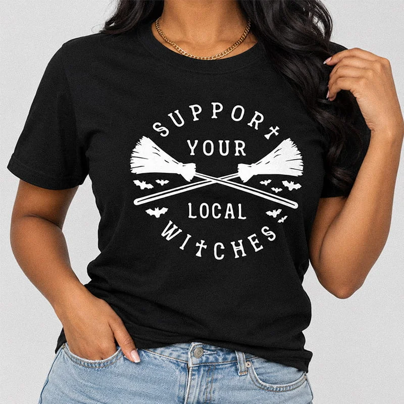 Woman wearing a black Support Your Local Witches Shirt - Witchy Halloween Tee featuring crossed broomsticks encircled by flying bats