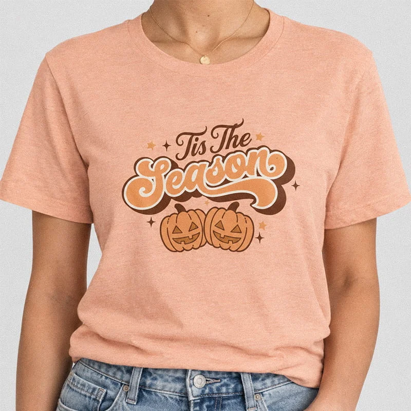 Woman wearing a heather peach Tis the Season Pumpkin T-Shirt - Halloween Fall Graphic Tee with two smiling jack-o'-lantern pumpkins and wavy typography reading Tis The Season