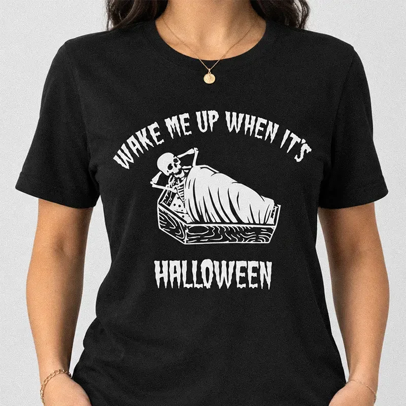 Woman wearing a black Wake Me Up When Its Halloween Shirt - Funny Skeleton Tee featuring a skeleton lying in a coffin graphic