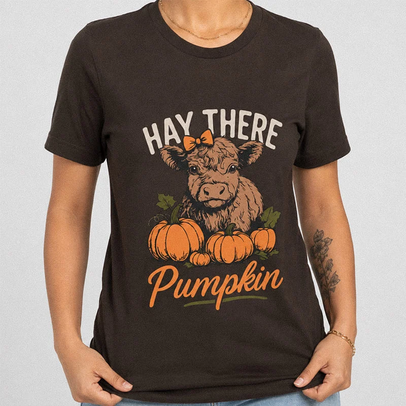 Woman wearing a dark brown Hay There Pumpkin Shirt - Cute Baby Highland Cow T-Shirt featuring a cute cow wearing a bow sitting among orange pumpkins