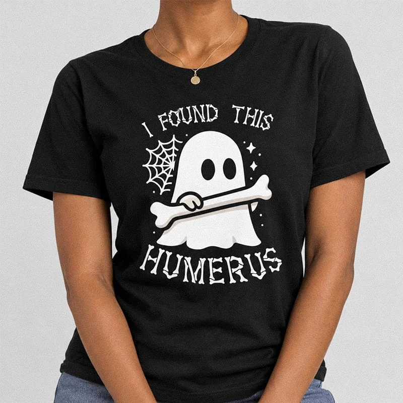 Woman wearing a black funny I Found This Humerus Shirt - Funny Humorous Ghost Tee featuring a cute ghost holding a bone next to a spider web with text reading I Found This Humerus