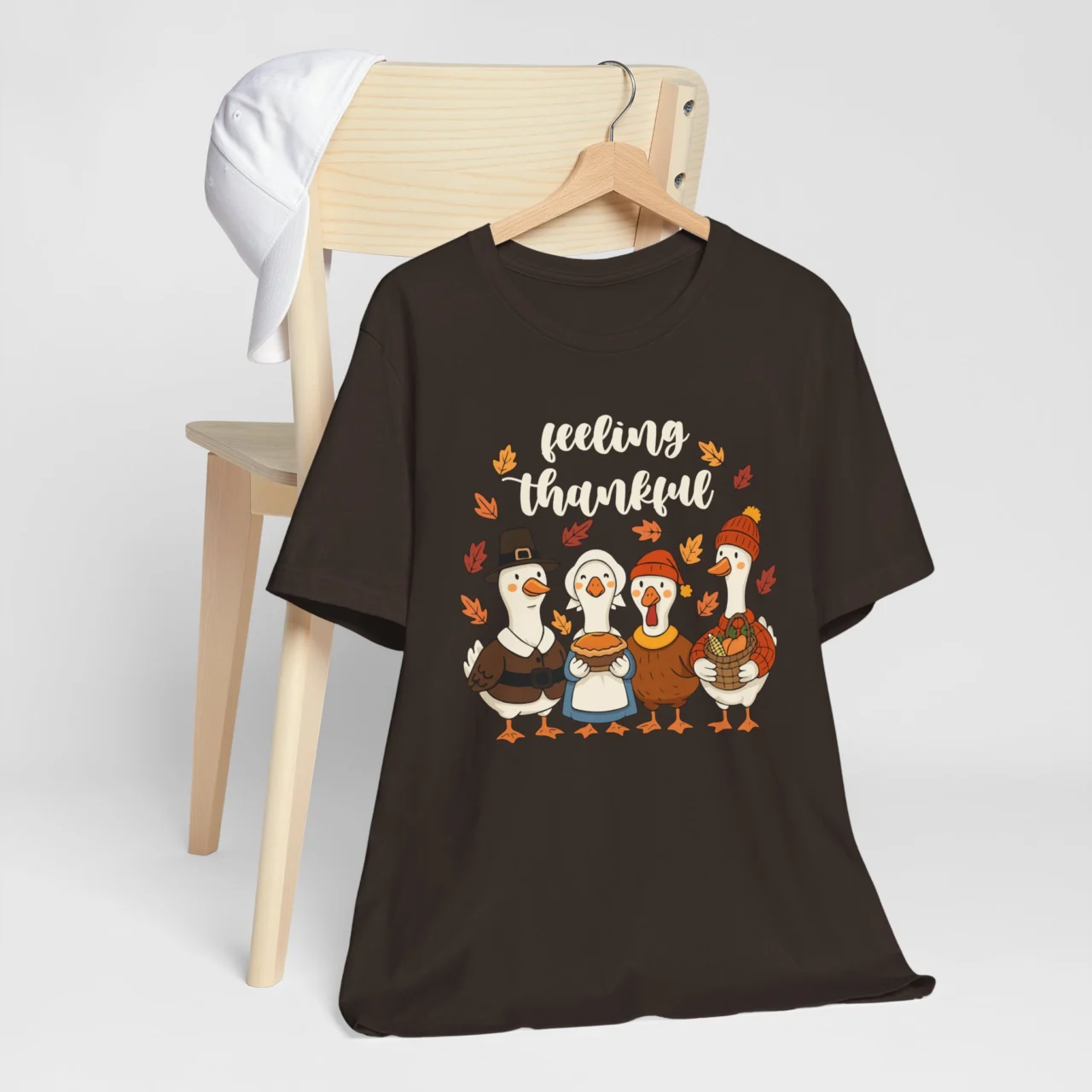 Black Feeling Thankful Geese Shirt with the text "Feeling Thankful" above four cartoon geese dressed in Thanksgiving pilgrim and festive clothes, surrounded by fall leaves, a cute holiday design.