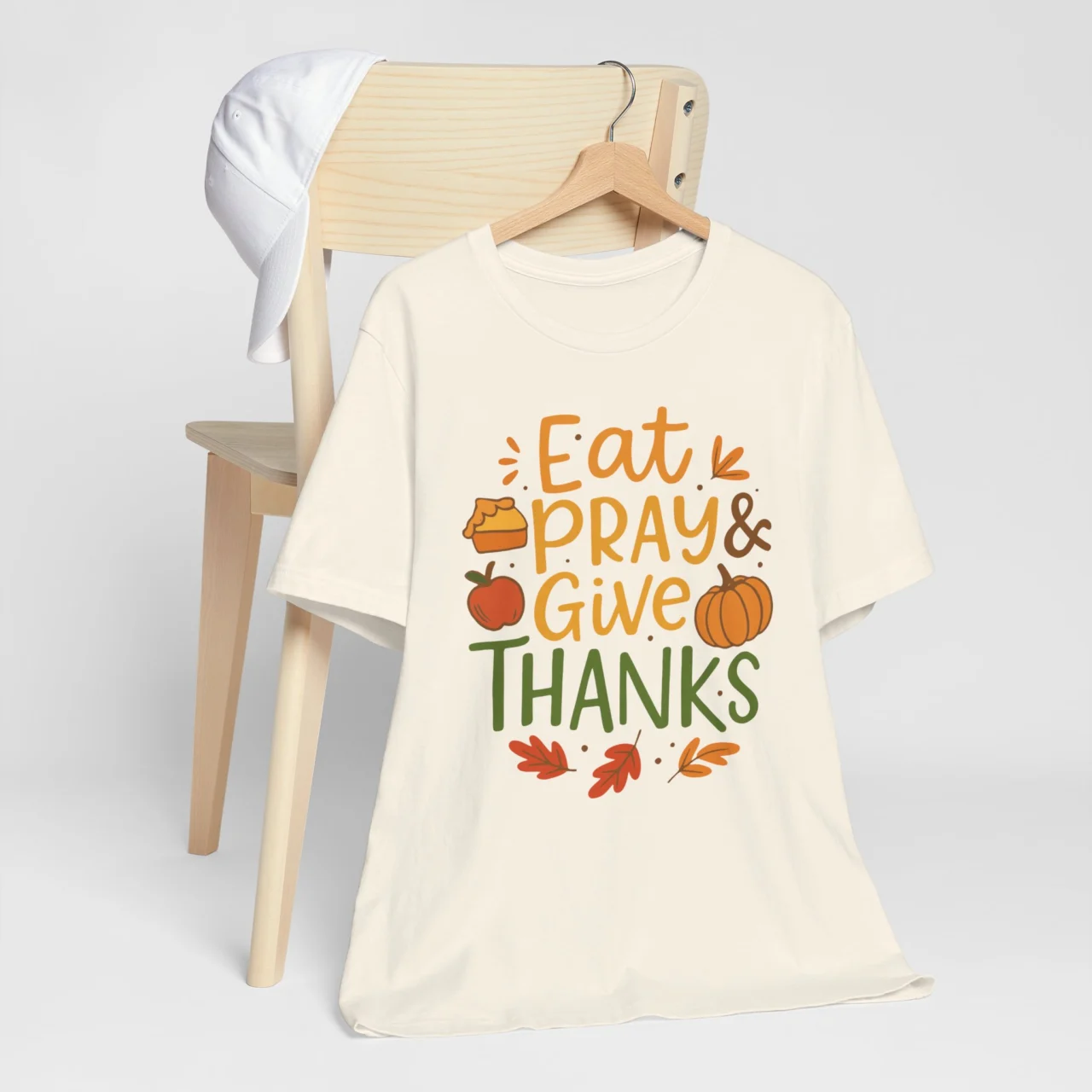 Natural Eat Pray Give Thanks Shirt with orange, green, and brown text saying "Eat Pray & Give Thanks" surrounded by illustrations of pie, an apple, a pumpkin, and fall leaves, a wholesome Thanksgiving design.