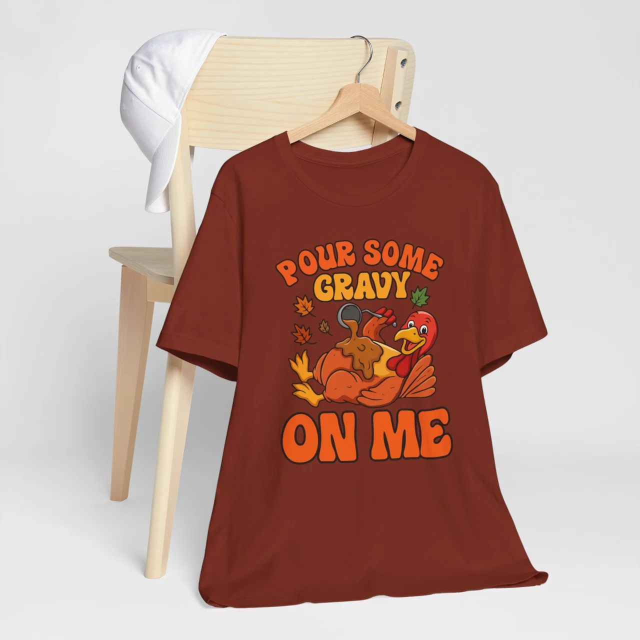 Pour Some Gravy On Me Turkey Shirt with a cartoon turkey being covered in brown gravy, with text on a maroon t-shirt, a funny sarcastic Thanksgiving design.
