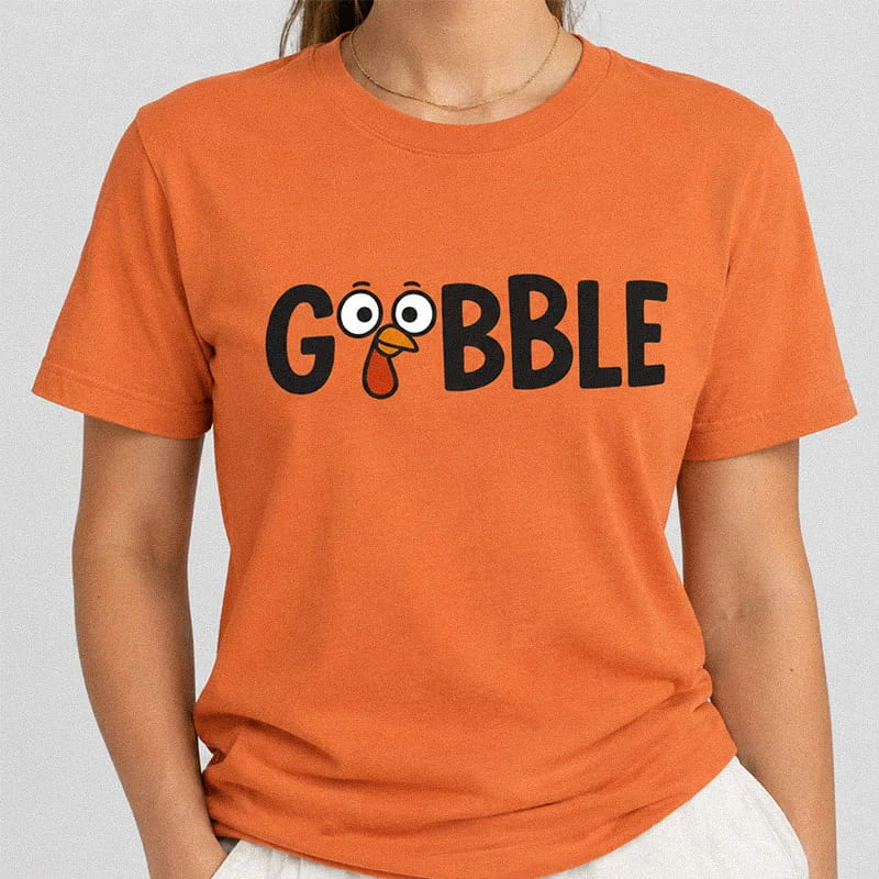 Woman wearing a burnt orange Funny Thanksgiving T-Shirt - Gobble Graphic Tee featuring the word GOBBLE with googly eyes and a turkey beak inside the Os